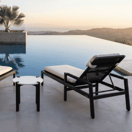 זווית אחורית של מיטת שיזוף מעוצבת MILOS מול הבריכה בשקיעה. ריהוט גן יוקרתי של LÜMA בגימור בזלת.